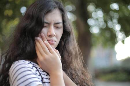 Young woman with long wavy brown hair and striped long-sleeved shirt holding her lower jaw with her face contorted in pain