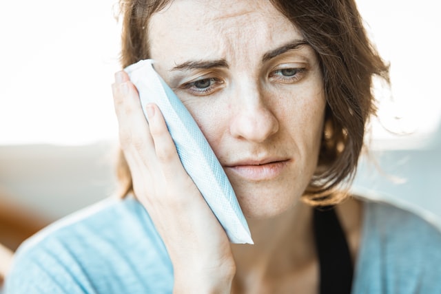 Woman with brown, shoulder-length hair cools her jaw with a cold pack and looks down sadly