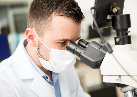 Man wearing a face mask and a white protective gown looks at something under a magnifying glass in a laboratory